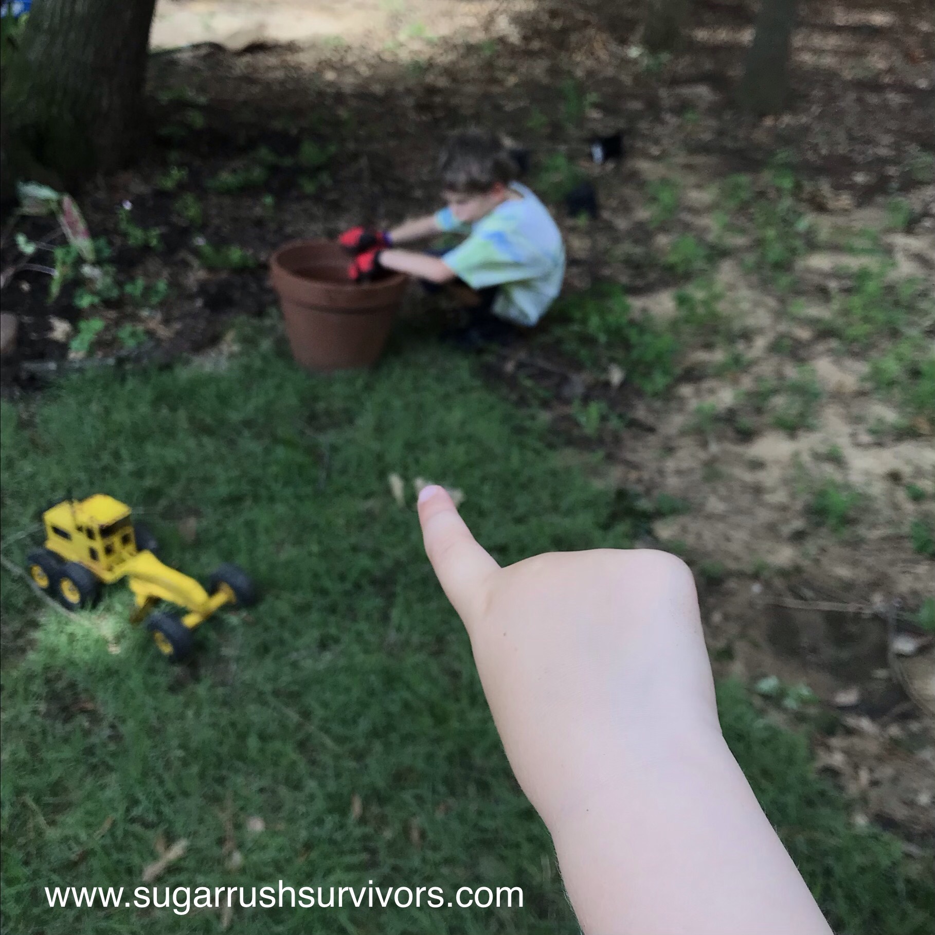 In the foreground a child's finger points to a young boy, blurred, in the background. The boy is crouched on the grass next to a large flowerpot. His posture is slouching and pouty. His arms rest on the flowerpot rim. A toy yellow tractor rests on the grass.